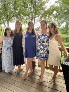 A group of five people standing on a wooden deck surrounded by trees. They are wearing summer dresses and smiling at the camera.