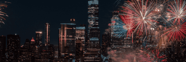 NYC skyline at night with tall, illuminated skyscrapers, including the Empire State Building, is shown. Colorful fireworks burst in red, orange, and blue, lighting up the sky.