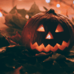 Three glowing jack-o’-lanterns with carved faces sitting on autumn leaves, surrounded by twinkling lights in the background.