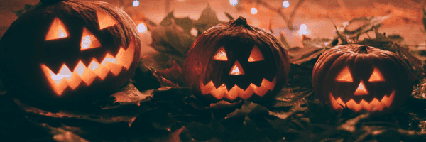 Three glowing jack-o’-lanterns with carved faces sitting on autumn leaves, surrounded by twinkling lights in the background.