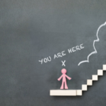 A conceptual image featuring a hand drawing a ladder with chalk on a blackboard, leading from the words "You are here" with a mark at the base, up to "Your Goal" at the top, indicating a path to achieve a goal.