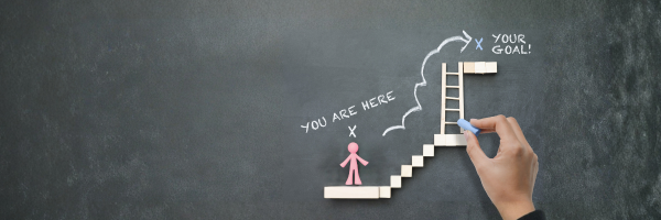 A conceptual image featuring a hand drawing a ladder with chalk on a blackboard, leading from the words "You are here" with a mark at the base, up to "Your Goal" at the top, indicating a path to achieve a goal.