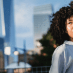 A person with curly hair smiles while talking on a phone and holding a cup. They are standing in an outdoor setting with trees and skyscrapers, including One World Trade Center, visible in the background.