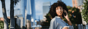 A person with curly hair smiles while talking on a phone and holding a cup. They are standing in an outdoor setting with trees and skyscrapers, including One World Trade Center, visible in the background.