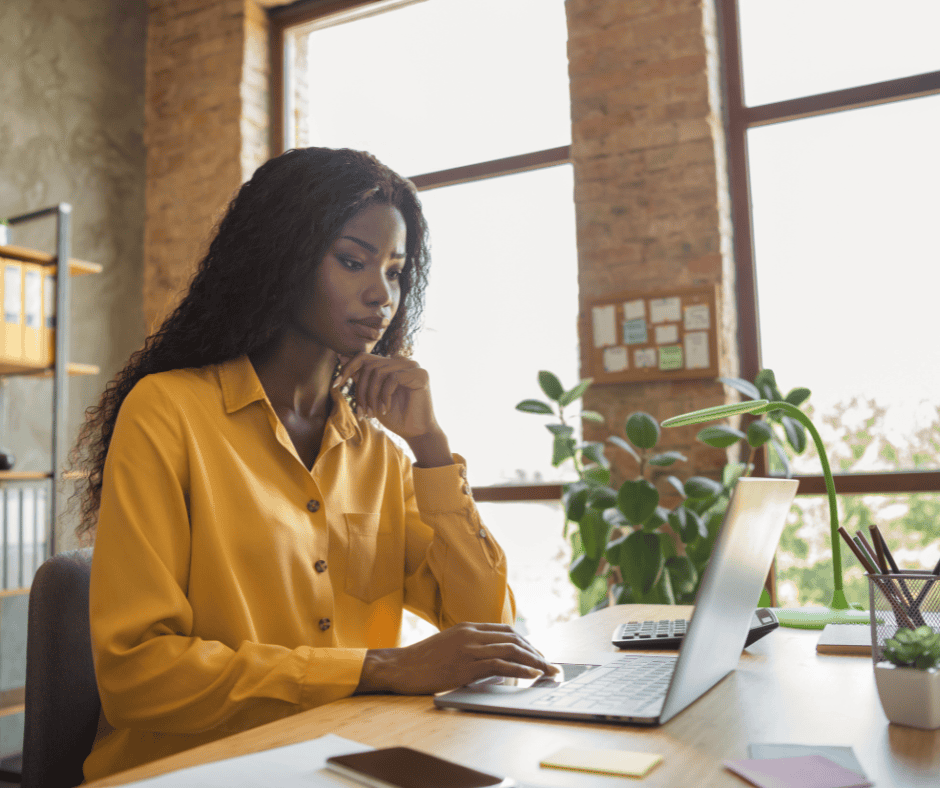 A person in a yellow shirt is working on a laptop at a desk in a bright office with large windows.