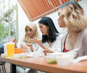 A group of people sitting at an outdoor table, enjoying food together. There are cups and plates with various dishes, along with a large yellow drink. The setting appears to be casual and friendly.