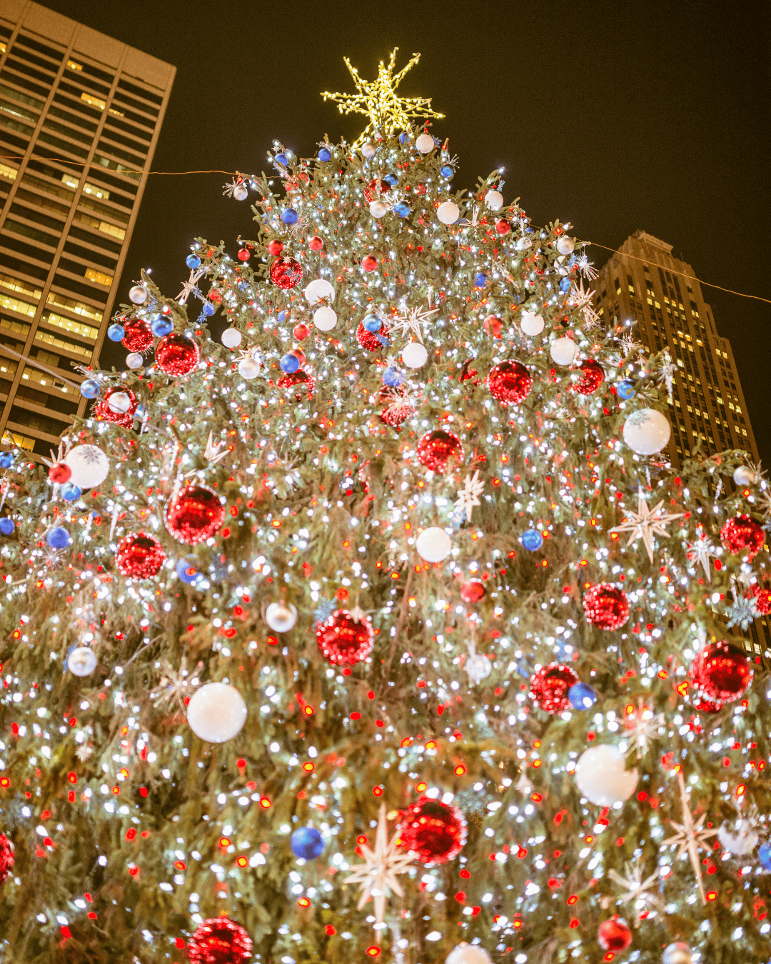 Close-up view of a brightly lit Christmas tree in New York City decorated with red, white, and blue ornaments, sparkling lights, and a glowing star topper against the night sky.