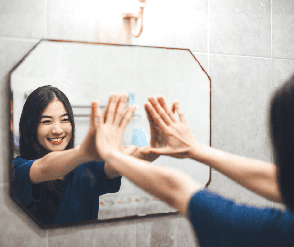 Person in a blue shirt smiling and making a heart shape with their hands, reflected in a bathroom mirror.