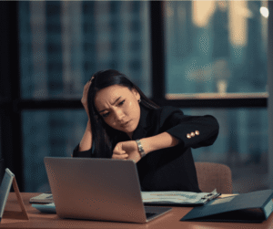 A person sits at a desk in an office, looking at their watch with a concerned expression. An open laptop and documents are on the desk, with a cityscape visible through the window.