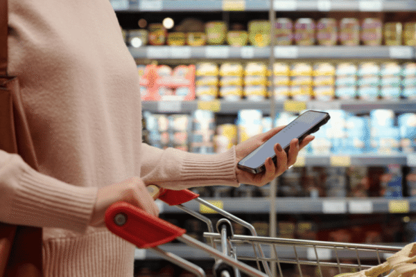 A person in a store aisle is holding a smartphone while pushing a shopping cart. Shelves stocked with various canned and packaged goods are visible in the background.