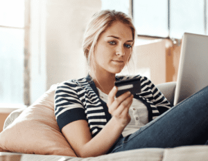 A person is sitting comfortably on a couch, holding a credit card and looking at a laptop. The setting is a well-lit, relaxed living space.