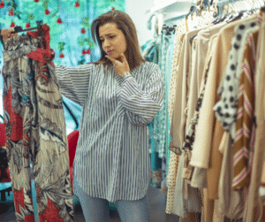 A person in a striped shirt is thoughtfully inspecting a pair of patterned pants in a clothing store. The store is filled with various garments hanging on racks, and there's a window in the background decorated with red hanging ornaments.