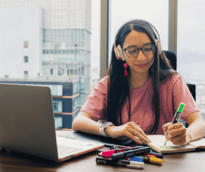 A person wearing headphones sits at a desk with a laptop, writing with a green marker. Various markers and a notebook are on the desk. A large window in the background shows a view of city buildings.