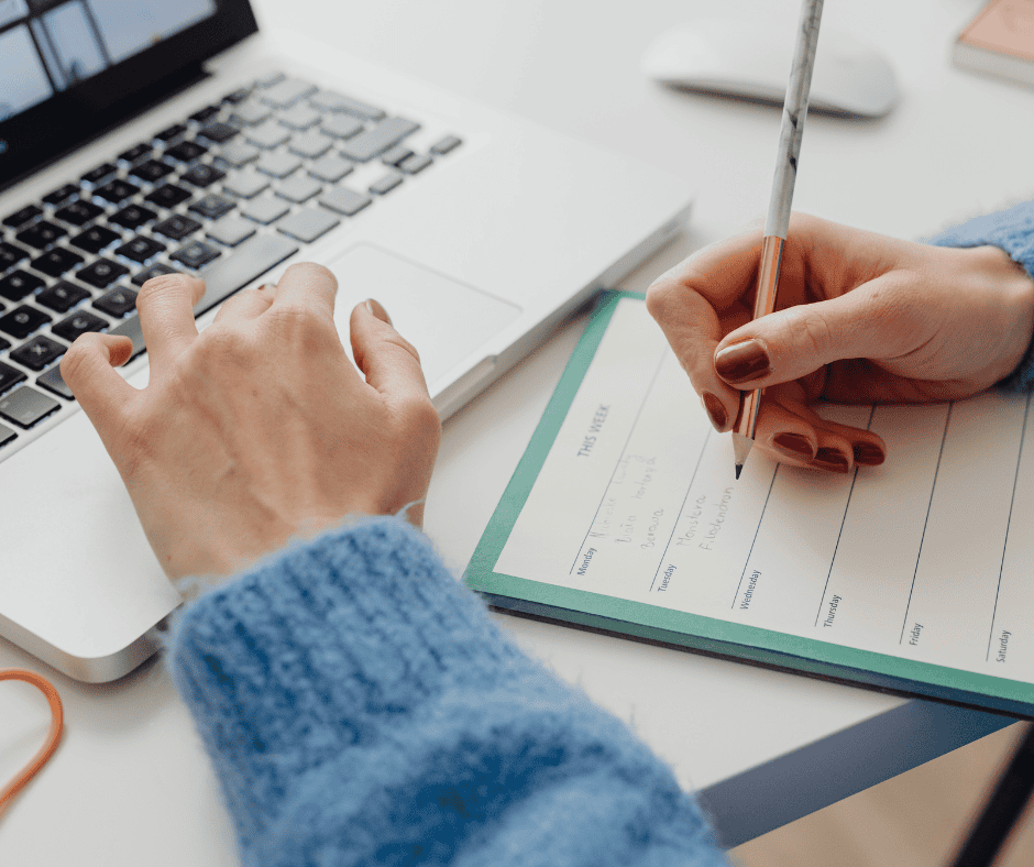 woman writing in a weekly planner while working on her laptop, organizing her schedule for a productive week ahead