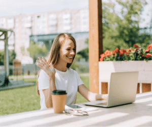 a person smiling and waving at a laptop outdoors, with a coffee cup nearby.