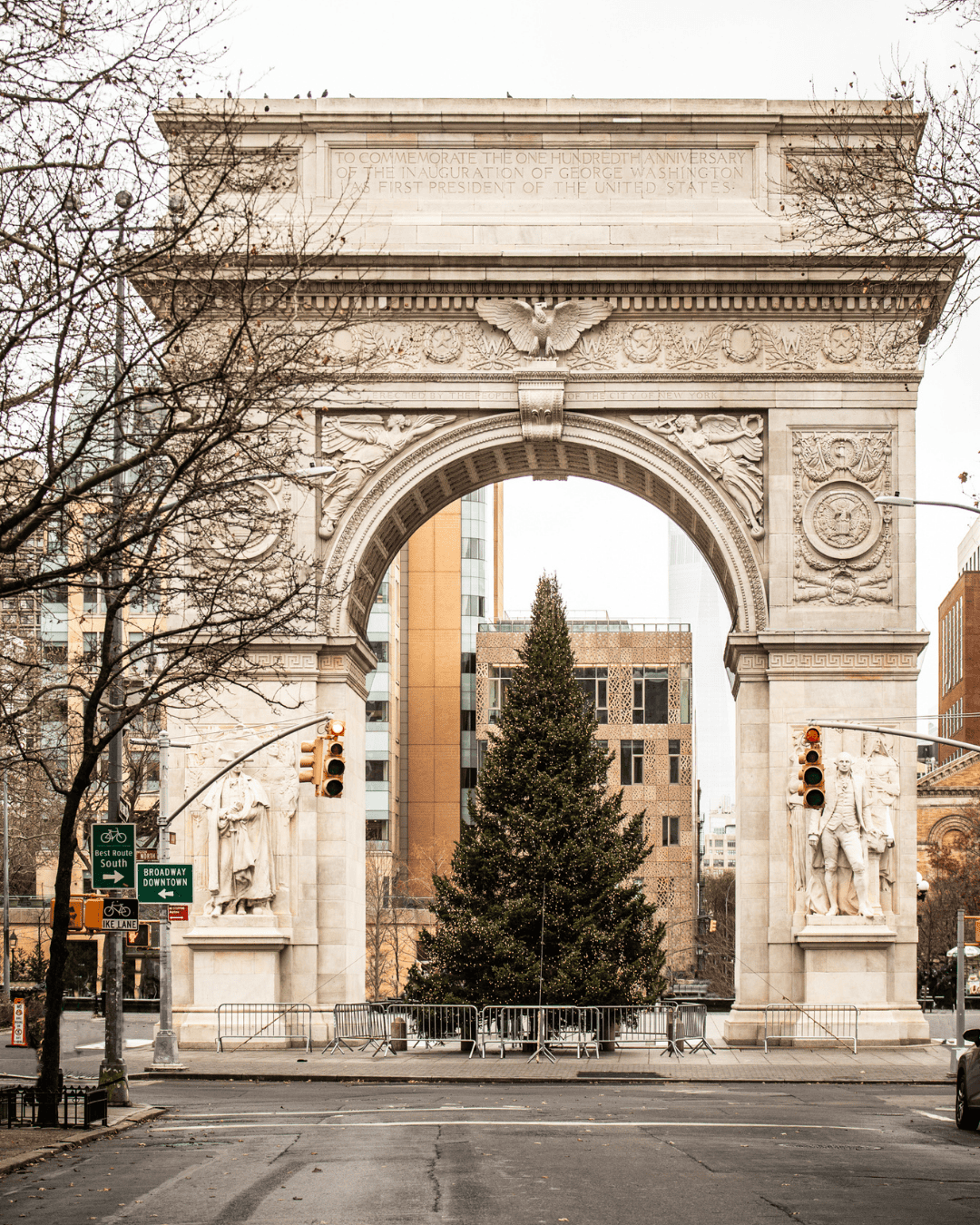 The Washington Square Park arch in New York City with a large Christmas tree centered beneath it, surrounded by winter trees and city buildings.