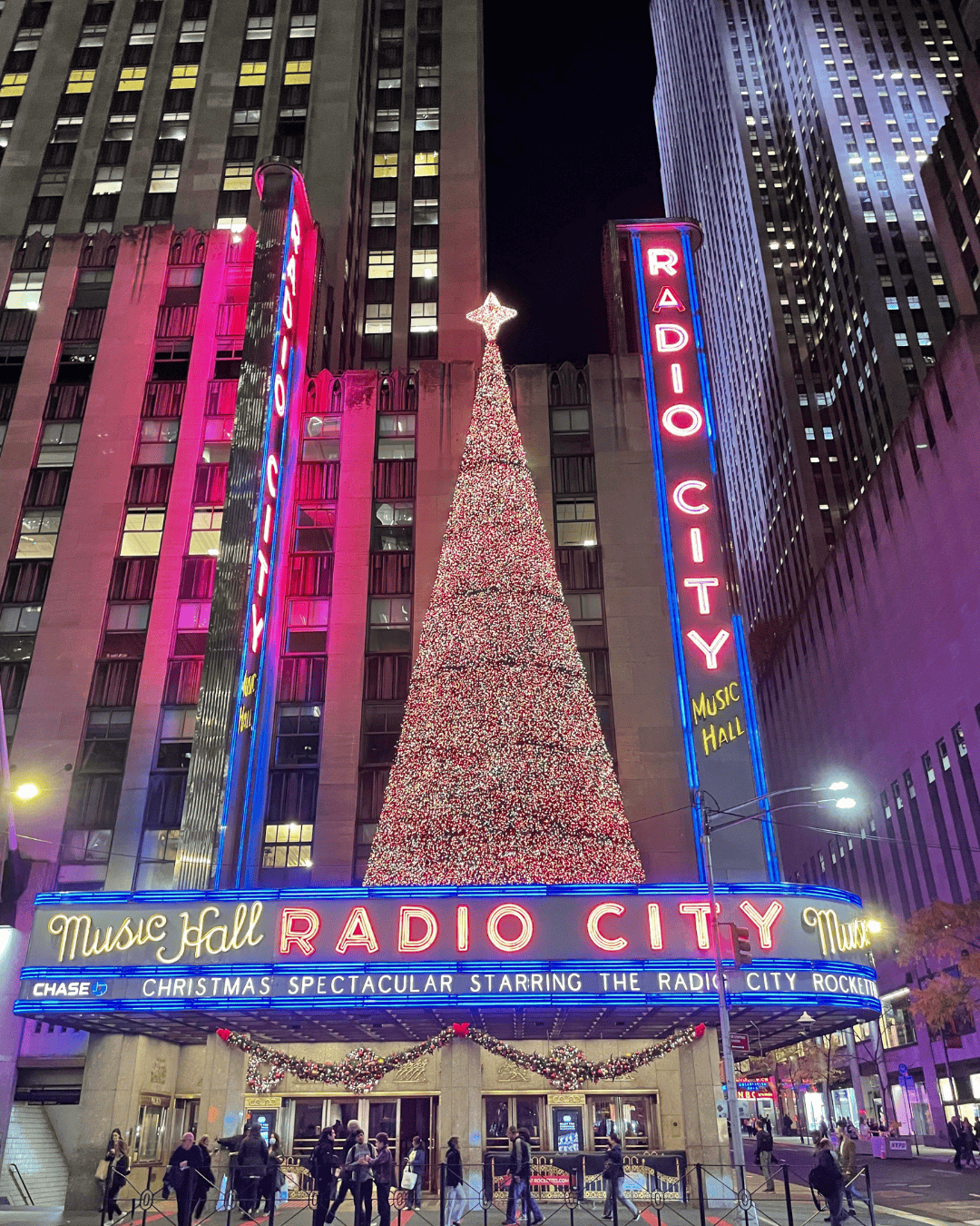 A large Christmas tree covered in colorful lights standing in front of a tall office building at night in New York City, with a golden statue in the foreground.