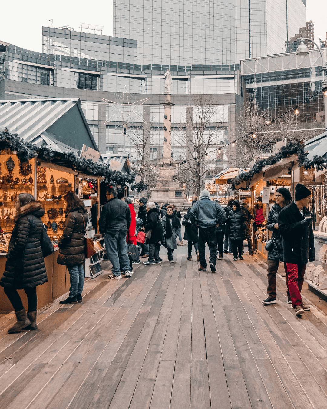People browsing festive holiday market stalls decorated with garland and lights in New York City, with winter coats, wooden walkways, and tall modern buildings in the background.