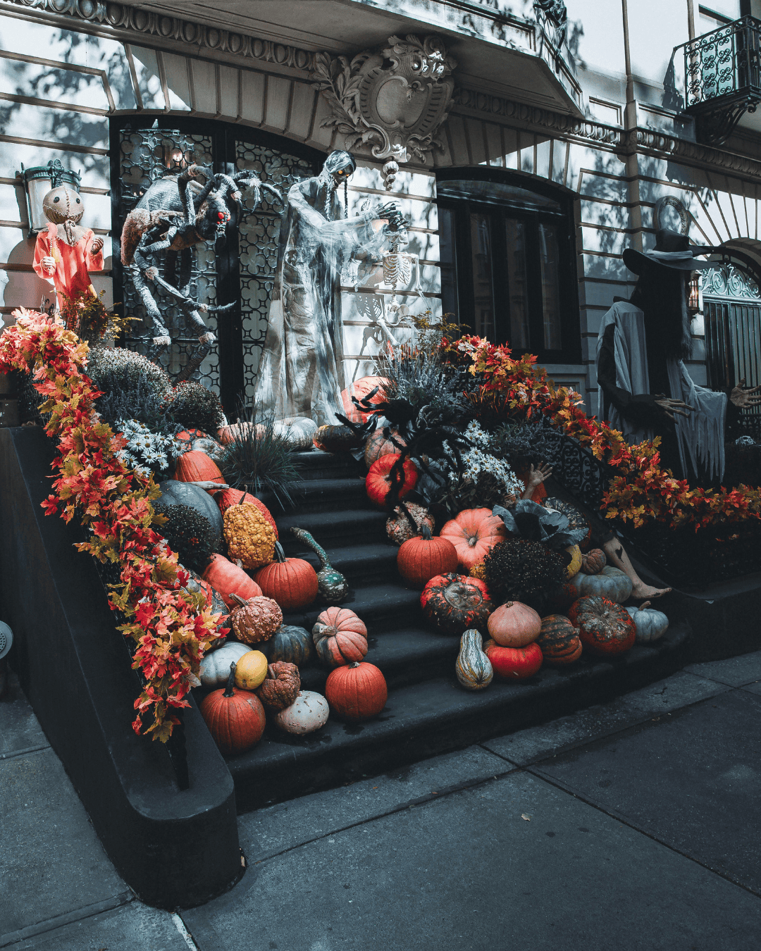 Brownstone steps in New York City decorated for Halloween with pumpkins, colorful autumn leaves, giant spiders, skeletons, and spooky life-sized figures.