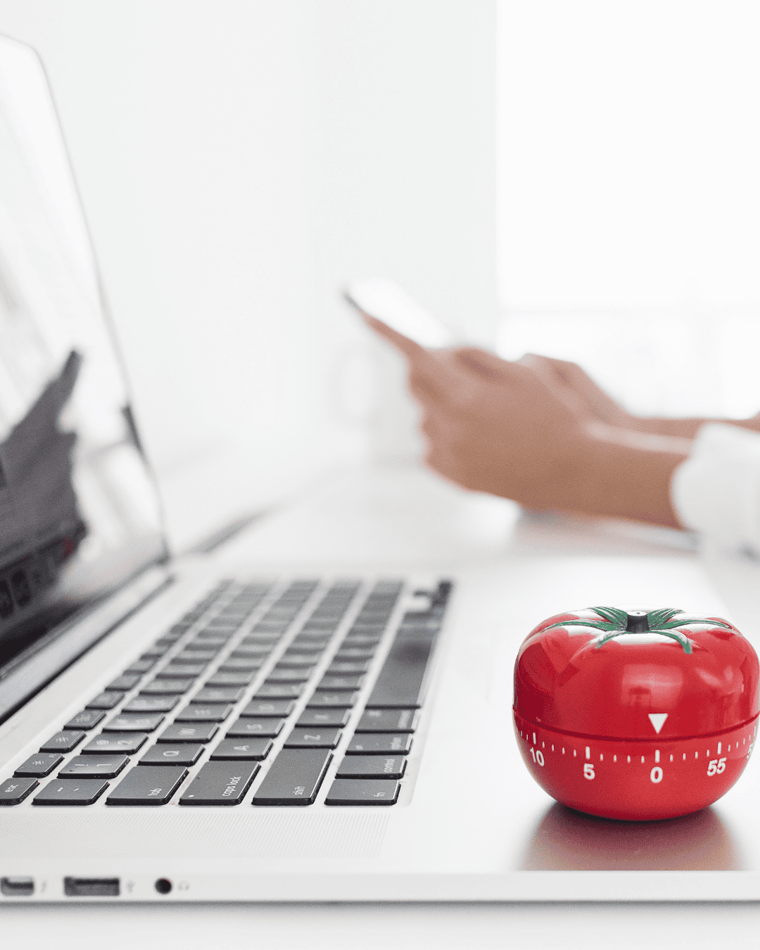 Close-up of a laptop with a red tomato-shaped kitchen timer on the keyboard, symbolizing the Pomodoro study method, with a person using a smartphone in the background.