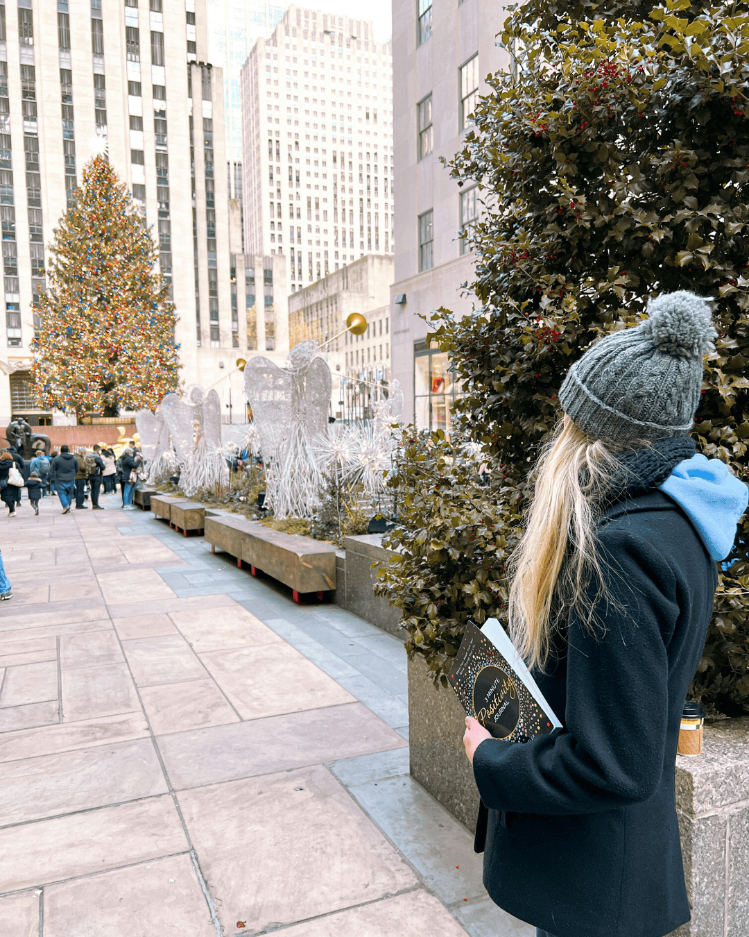 Woman in a winter coat and knit hat standing near holiday decorations at Rockefeller Center, looking toward the large Christmas tree and angel sculptures in New York City.