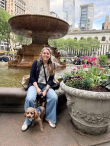 A person sits on the edge of a large fountain in Bryant Park, New York City. They have a small dog beside them on a leash. Behind them are skyscrapers and lush greenery. A stone planter with vibrant flowers is in the foreground.