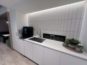 Image of a modern kitchen styled as a coffee station with a sign reading "Webb's Coffee Station." The kitchen features a sink, espresso machine, and potted plants, with white countertops and gray tile backsplash.