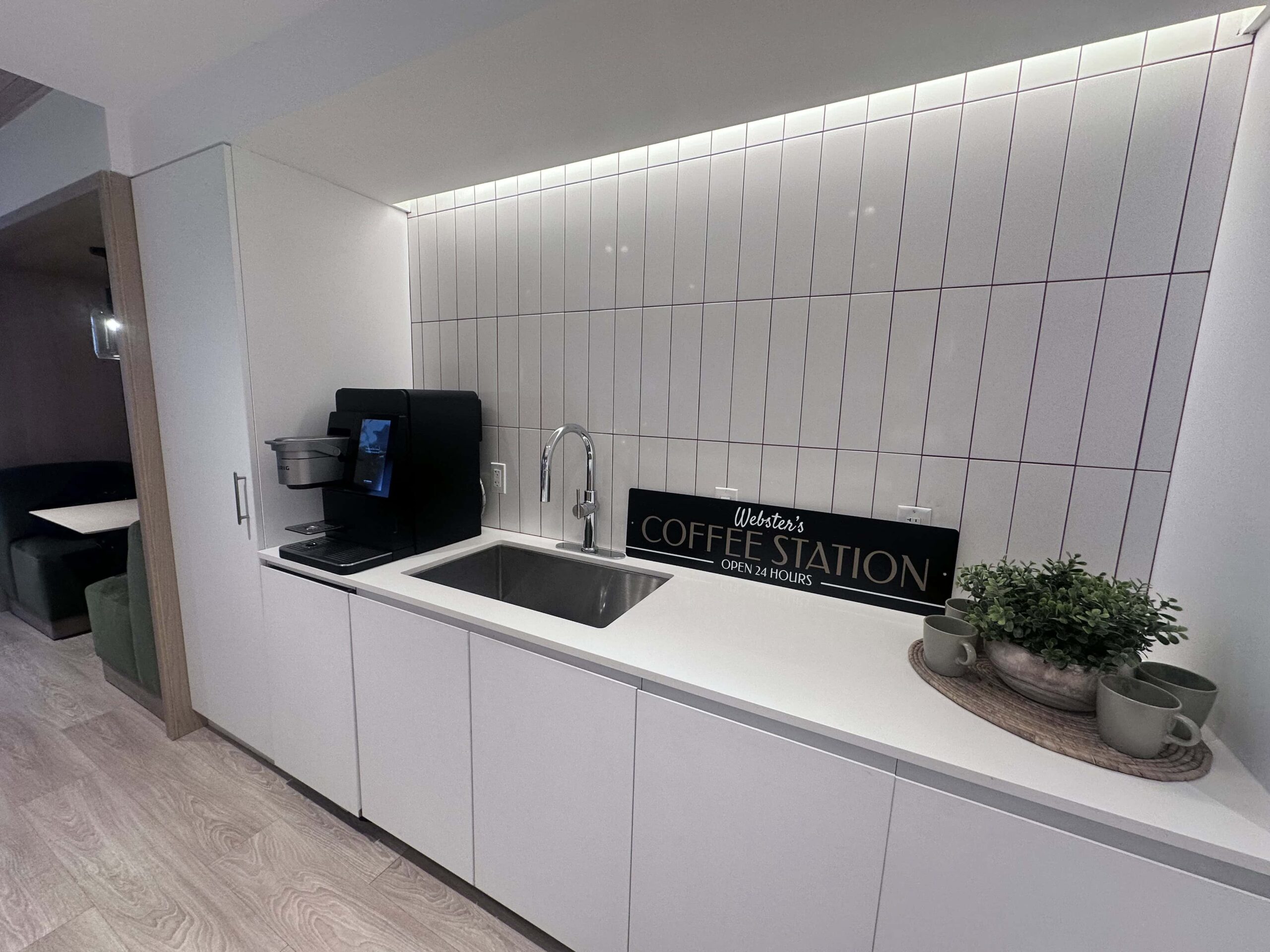 Image of a modern kitchen styled as a coffee station with a sign reading "Webb's Coffee Station." The kitchen features a sink, espresso machine, and potted plants, with white countertops and gray tile backsplash.