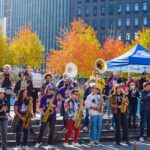 A lively outdoor band performance during the NYC Marathon at the Plaza at 300 Ashland in Brooklyn. Musicians play brass and woodwind instruments under a bright blue tent, surrounded by colorful autumn trees and a cheerful crowd enjoying the event.