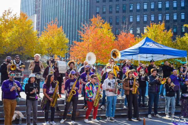A lively outdoor band performance during the NYC Marathon at the Plaza at 300 Ashland in Brooklyn. Musicians play brass and woodwind instruments under a bright blue tent, surrounded by colorful autumn trees and a cheerful crowd enjoying the event.