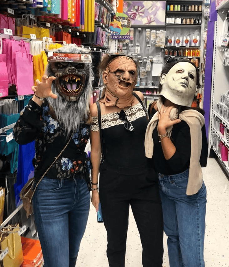 Three friends in a party store trying on Halloween masks—a werewolf, a horror character, and Michael Myers—while posing playfully in the aisle.