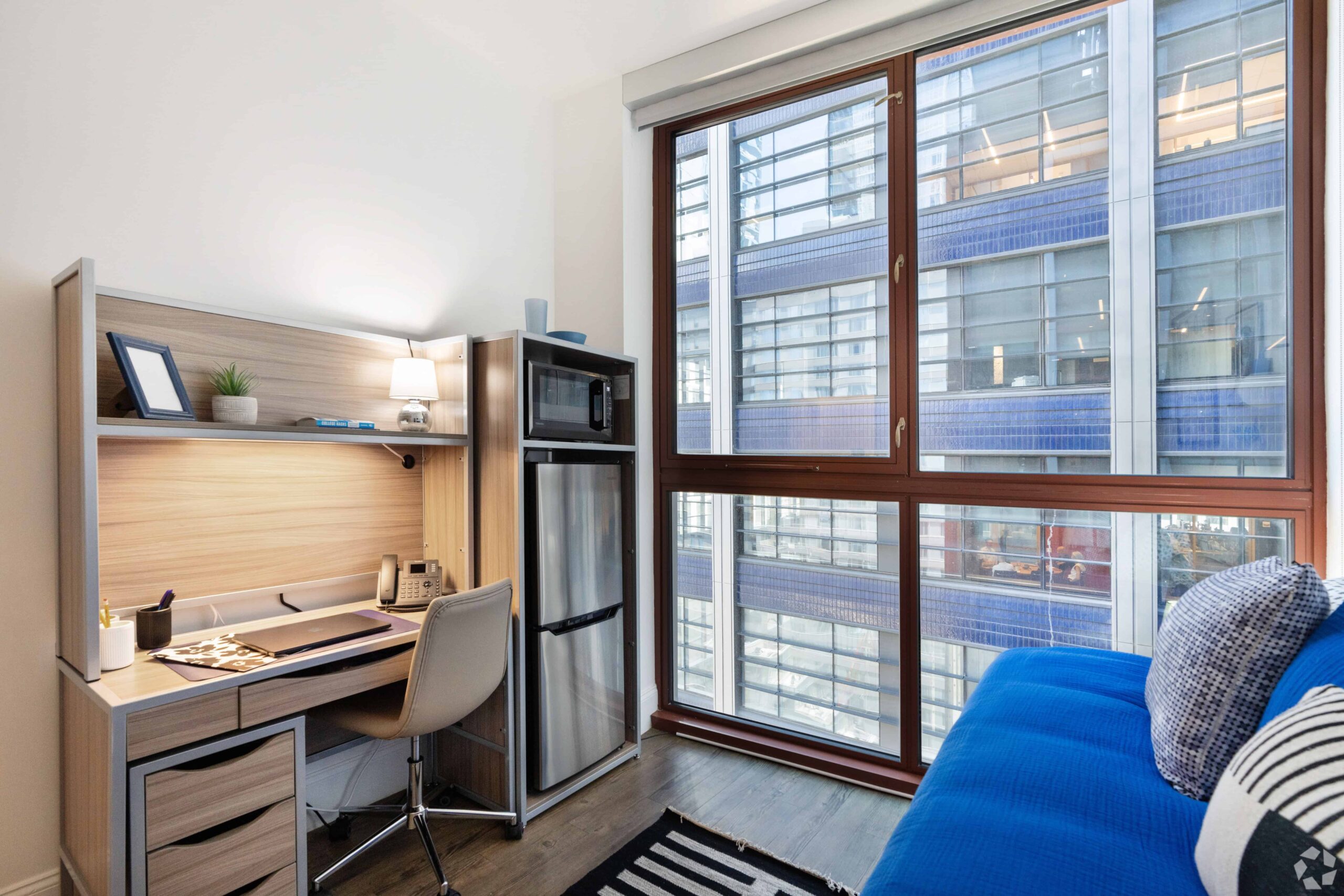 Modern studio apartment interior featuring a desk with shelves, a mini-fridge, a large window overlooking an urban environment, and a blue sofa with cushions.