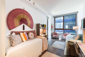 Interior of a modern apartment featuring a bedroom with a large bed and decorative fan on the wall, a living area with stylish chairs and a study area with a desk and bookshelf. Large windows offer a view of city buildings outside.