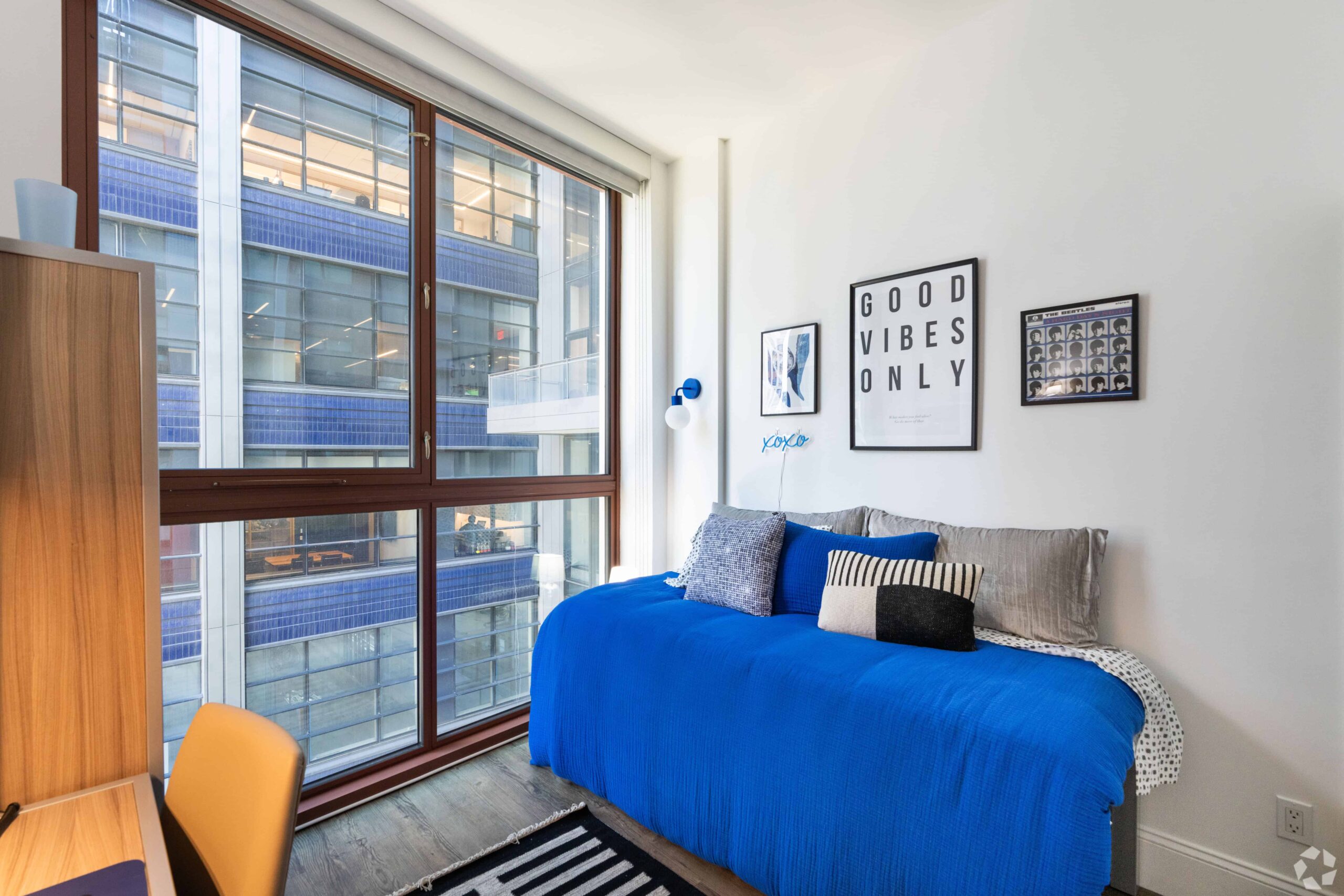 Alt text: A bright and modern furnished room at Webster Apartments featuring a large window with city views, a twin bed with a bold blue comforter and decorative pillows, wall art including a “Good Vibes Only” print, and a small desk and chair in the corner.