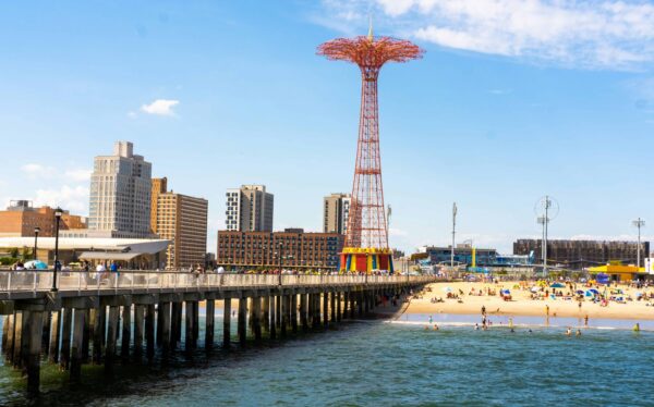 A beach scene at Coney Island with a pier extending into the water. The Parachute Jump tower is prominently visible. People are enjoying the beach under a clear blue sky, and various buildings line the shoreline in the background.