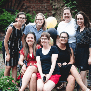 A group of people are smiling and posing with copies of the book "Then She Was Gone" on a rooftop with city buildings in the background. Some hold drinks, and the scene is lit warmly.
