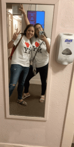Two individuals pose in front of a mirror wearing matching "I ❤️ NY" shirts. One person is making a peace sign, and the other is holding up a smartphone. A sanitizer dispenser is mounted on the wall beside them.