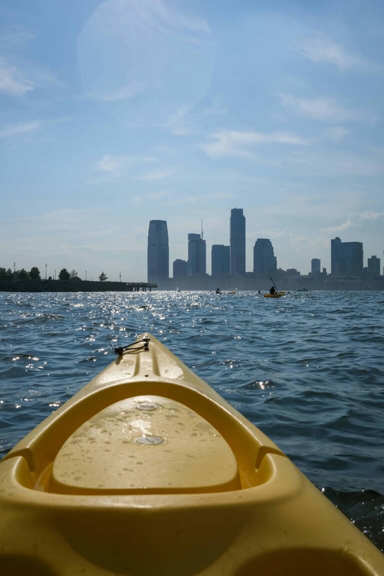 A yellow kayak floats on the water with a view of the New York City skyline in the background. The sun glimmers on the surface of the water, and other kayakers are visible in the distance.